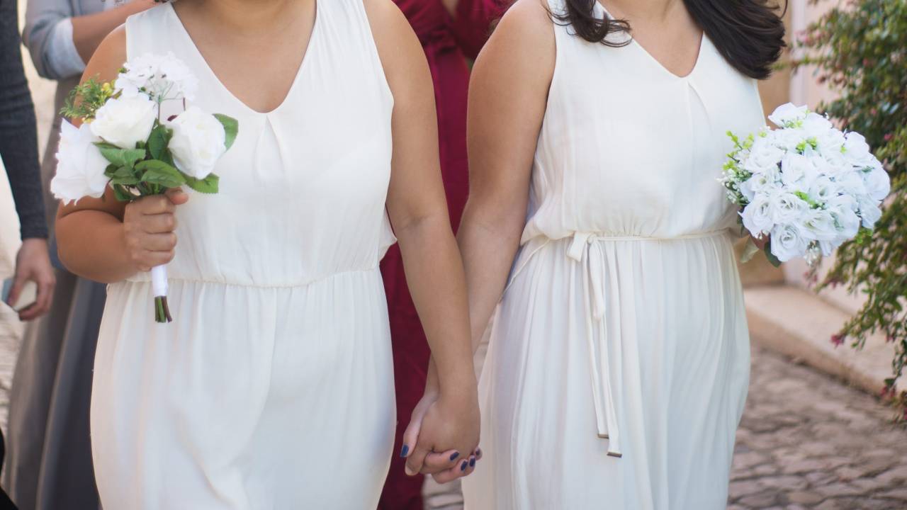 Foto mostra uma mulher negra e uma mulher branca de mãos dadas casando, cada uma usando vestido branco e buquê de rosas brancas