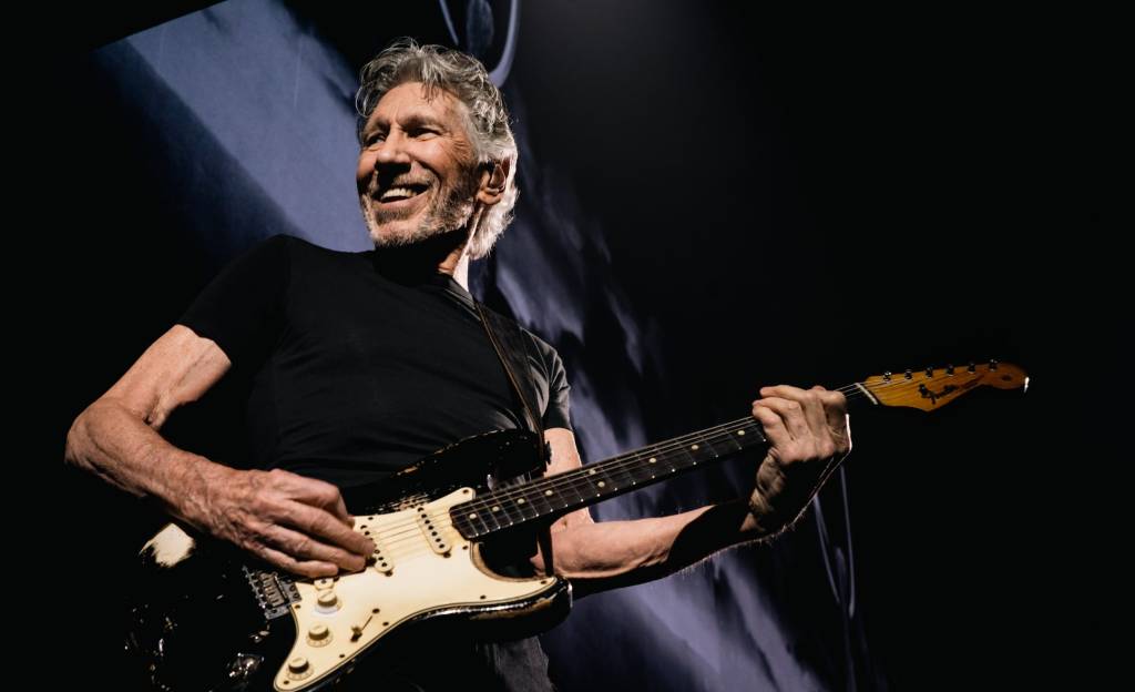 Roger Waters é um homem branco de cabelo grisalho. Ele usa blusa preta e está tocando uma guitarra Fender preta e branca