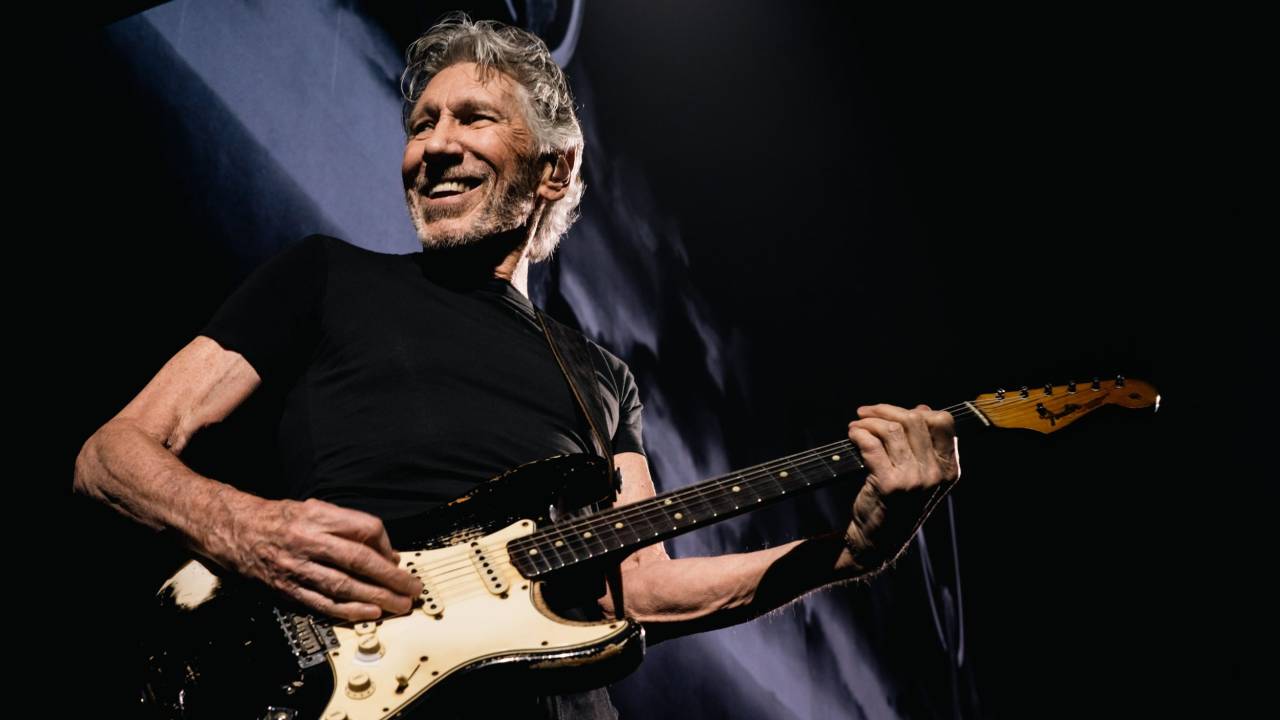 Roger Waters é um homem branco de cabelo grisalho. Ele usa blusa preta e está tocando uma guitarra Fender preta e branca