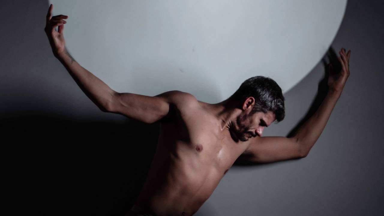 Thiago Soares está sem camisa e segura uma esfera branca nas costas, em foto com luz e sombra