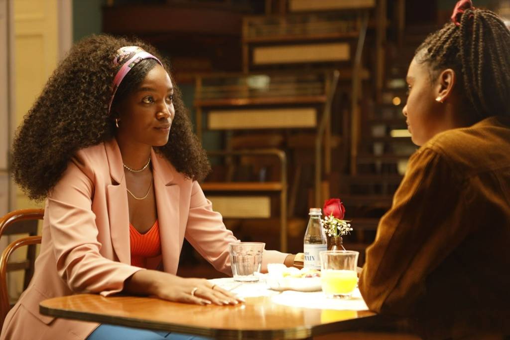 Iza e Gabz são duas mulheres negras. Elas estão sentadas, cada uma em uma ponta da mesa, Iza de frente para a câmera. Iza usa top vermelho, blazer rosa claro e faixa rosa clara no cabelo. Gabz usa casaco marrom.