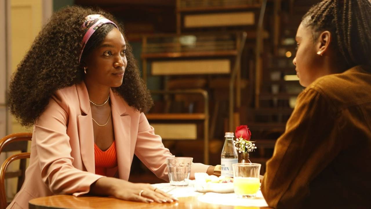 Iza e Gabz são duas mulheres negras. Elas estão sentadas, cada uma em uma ponta da mesa, Iza de frente para a câmera. Iza usa top vermelho, blazer rosa claro e faixa rosa clara no cabelo. Gabz usa casaco marrom.