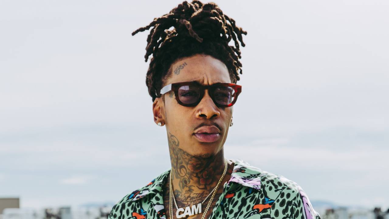 Wiz Khalifa é negro, está de camisa estampada, um colar dourado com a palavra CAM, usa o cabelo preso no alto da cabeça com dreads na ponta e óculos escuros