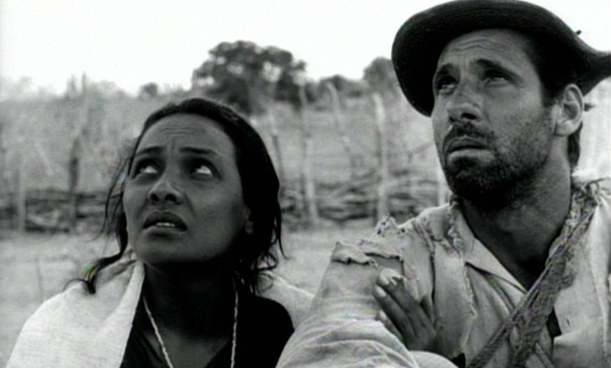 Foto em preto e branco da atriz Maria Ribeiro e do ator Átila Iorio caracterizado com os personagens de Vidas Secas, em meio a uma paisagem árida