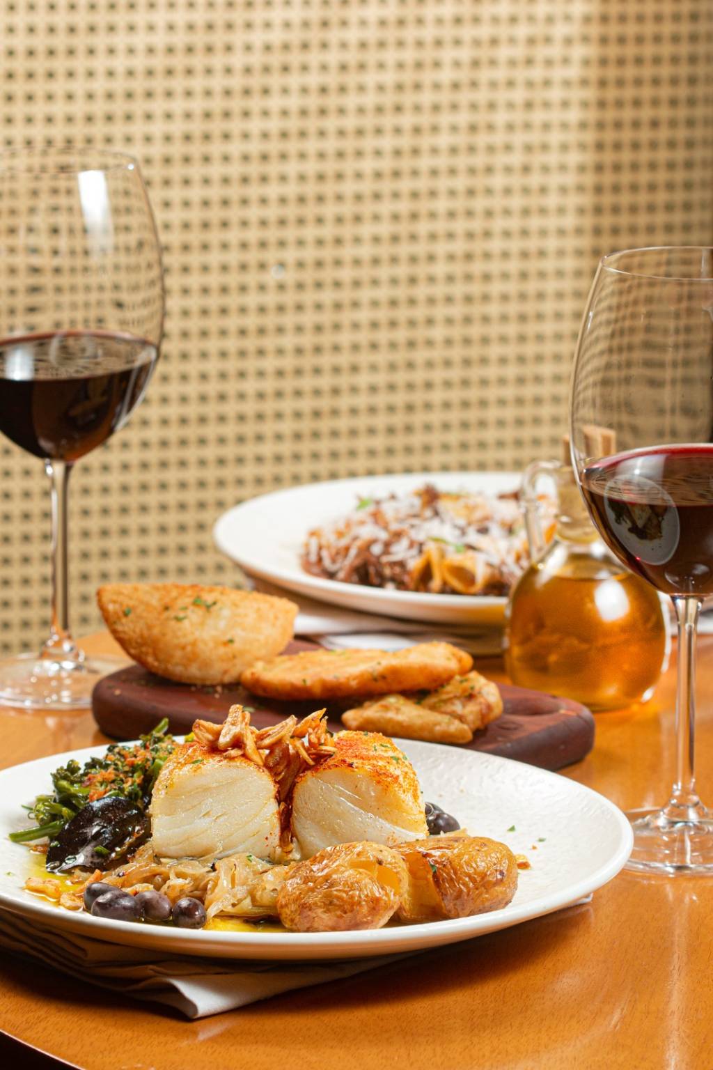 Foto mostra mesa com prato de bacalhau e duas taças de vinho tinto