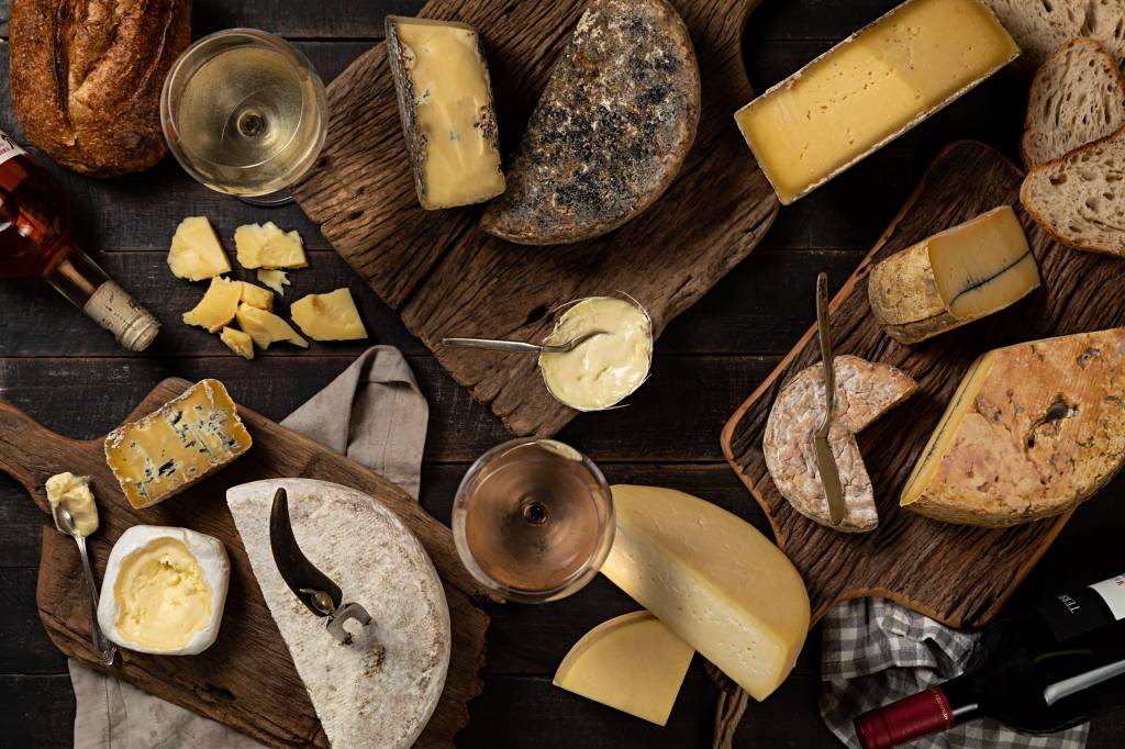 Mesa com diversos queijos, duas taças cheias e uma garrafa de vinho, fotografada de cima
