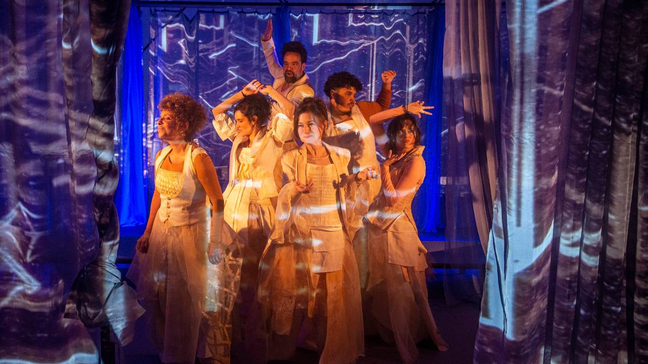Dois atores e quatro atrizes com roupas claras na Nave de Luz, uma estrutura com tecidos sobre os quais estão projetadas imagens em tons azulados.