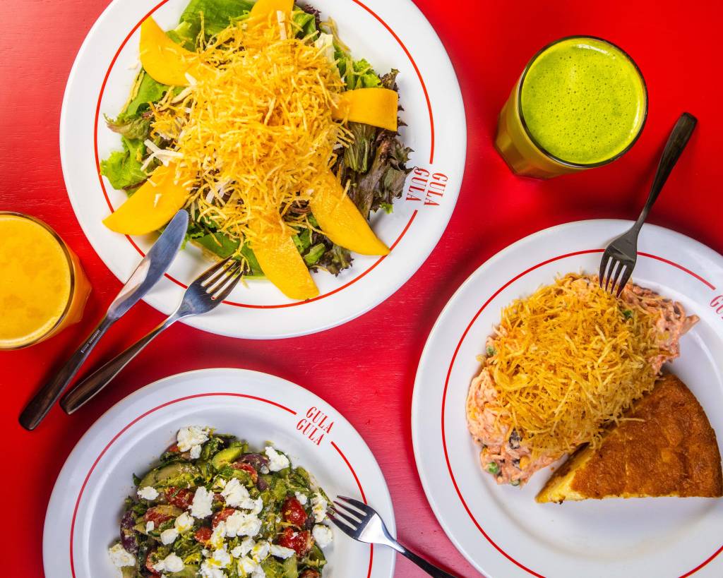 Mesa posta com toalha vermelha com uma prato de salada com manga e batata-palha, um prato de quiche com empadão com batata-palha, um prato com salada com queijo por cima e um copo de suco verde.