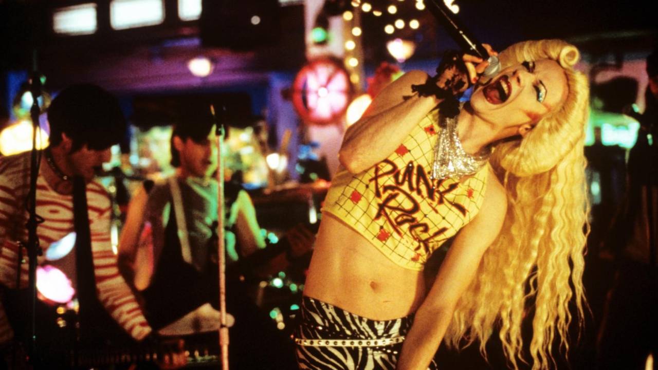 A drag Hedwig usa peruca loura platinada, muita maquiagem e veste calça com estampa de zembra e top cropped onde se lê punk rock.