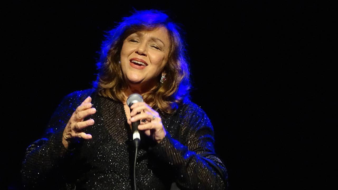 Jane Duboc está de olhos fechados e cantando ao microfone. Ela usa camisa preta com brilhos de manga comprida. Uma luz azul incide em seus cabelos, obros e brancos.