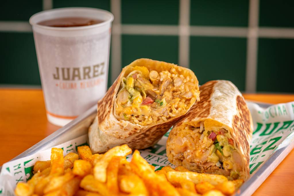 A foto tem um burrito cortado ao meio e batatas fritas. Ao fundo, um copo branco de papel com bebida onde se lê o nome Juarez.
