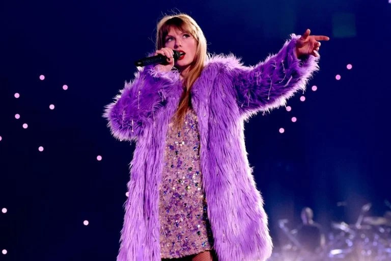 Foto mostra Taylow Swift usando casaco roxo de pelos e vestido prata brilhante em show