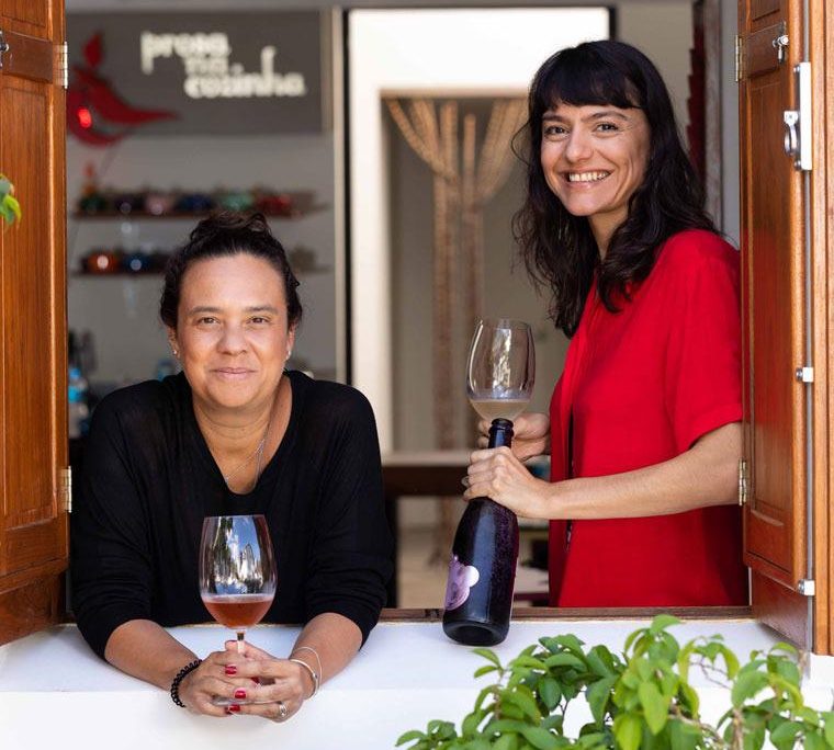 Foto mostra duas mulheres segurando taças de vinho em janela