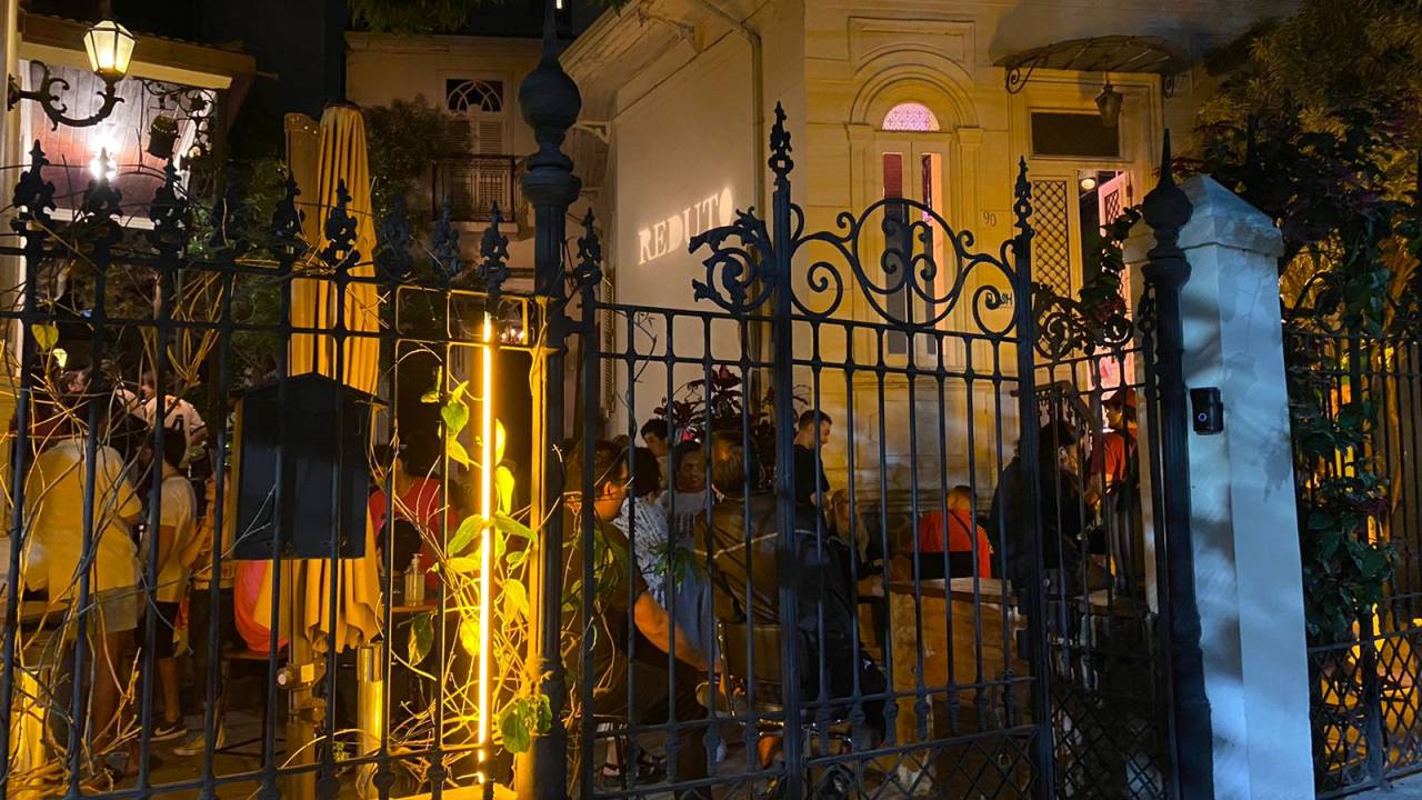 Foto à noite do portão de ferro do Reduto, com o casarão branco aparecendo à direita e pessoas em pé.