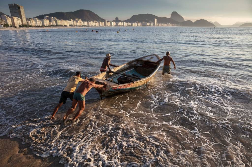 Quatro pescadores no posto 6 da Praia de Copacabana empurram um barco na areia da praia para o mar, tendo ao fundo o Morro da Urca e o Pão de Açúcar.