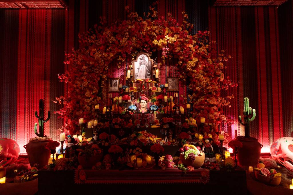 Com um fundo vermelho, imagem traz estante com diversas imagens de Frida Kahlo, frutas e velas