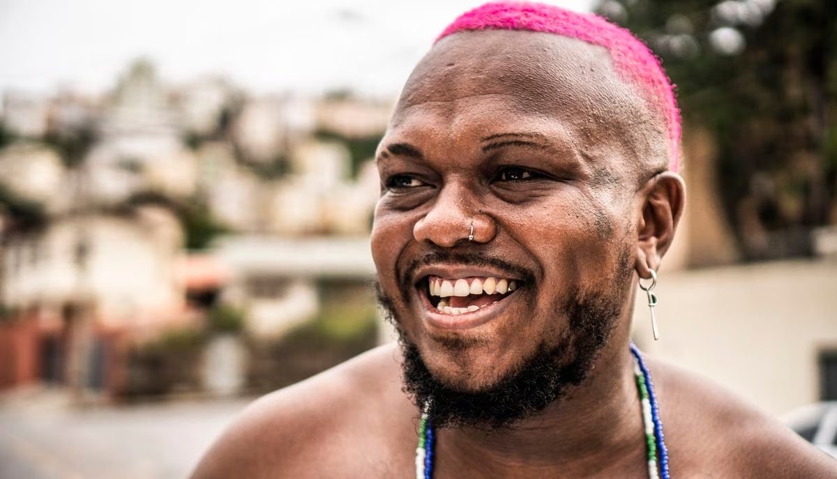 O rapper Djonga é um homem negro com cabelo curto pintado de rosa-choque. Ele está sem camisa, com guias de orixá no pescoço, olhando para o lado e sorrindo.