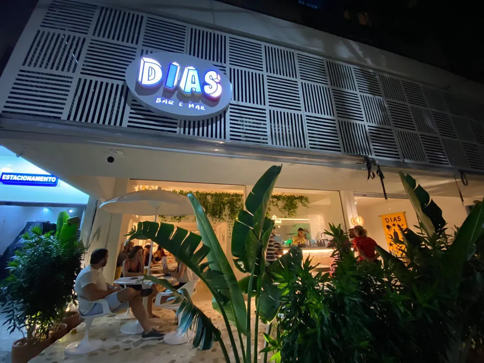 A fachada do Dias, com um letreiro branco com grafismos onde se lê Dias Bar e Mar, plantas na frente da loja e mesinhas brancas com pessoas sentadas nas cadeiras.