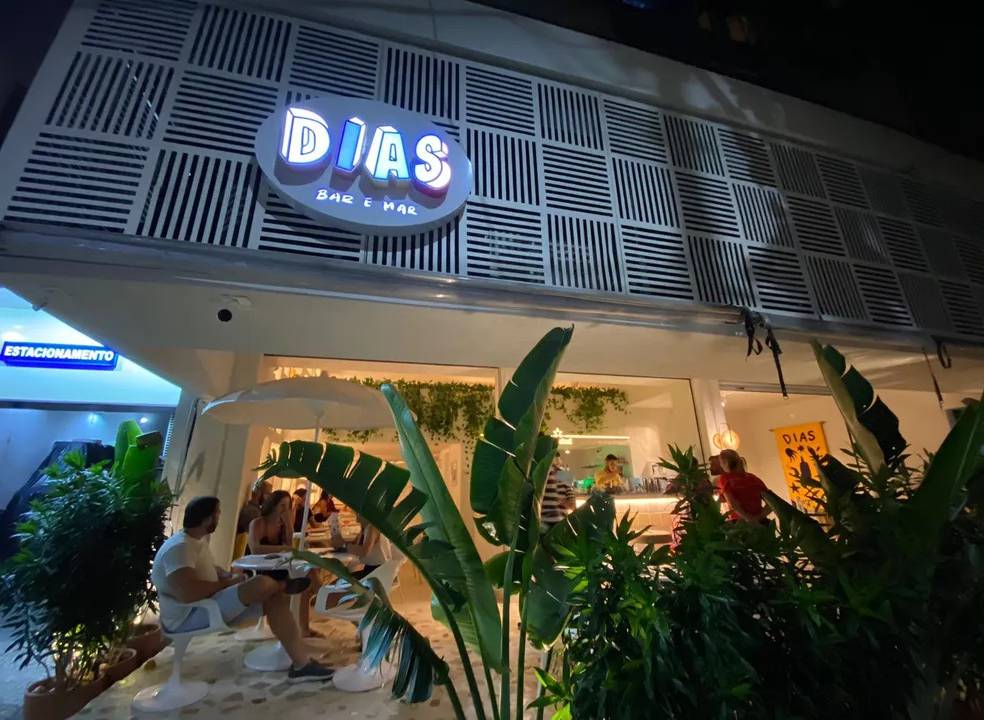 A fachada do Dias, com um letreiro branco com grafismos onde se lê Dias Bar e Mar, plantas na frente da loja e mesinhas brancas com pessoas sentadas nas cadeiras.