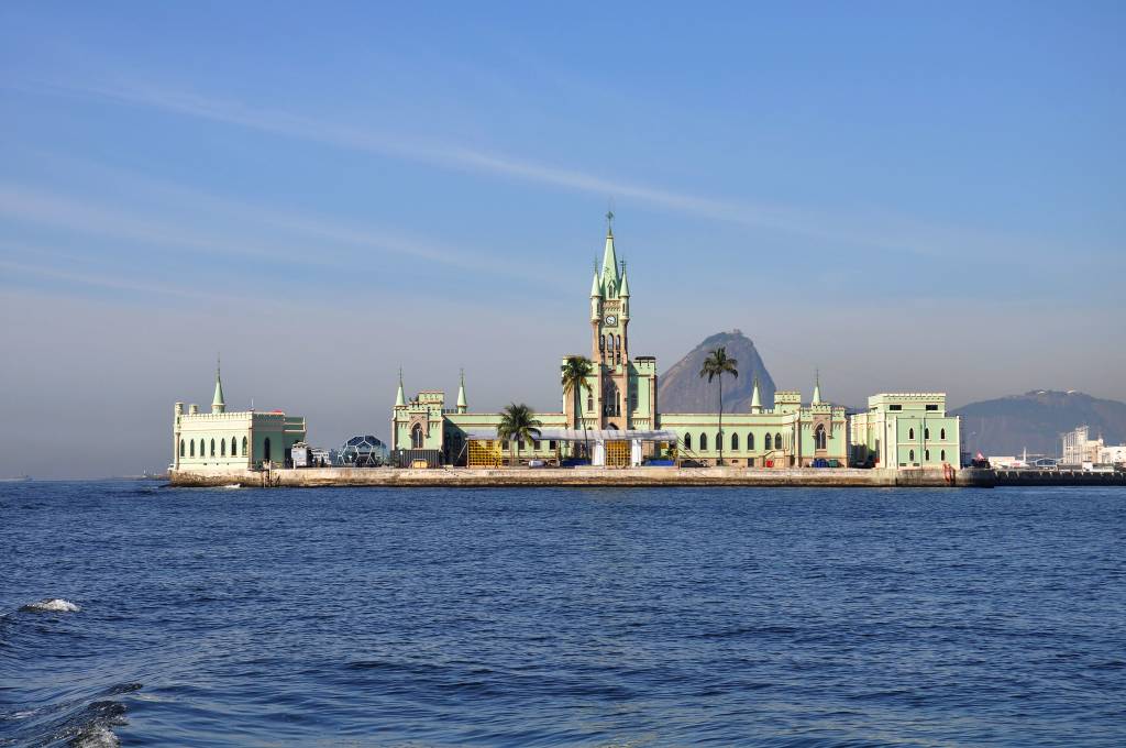 A Ilha Fiscal vista de longe, com o mar na frente, o palacete neogótico verde na filha e o Pão de Açúcar atrás