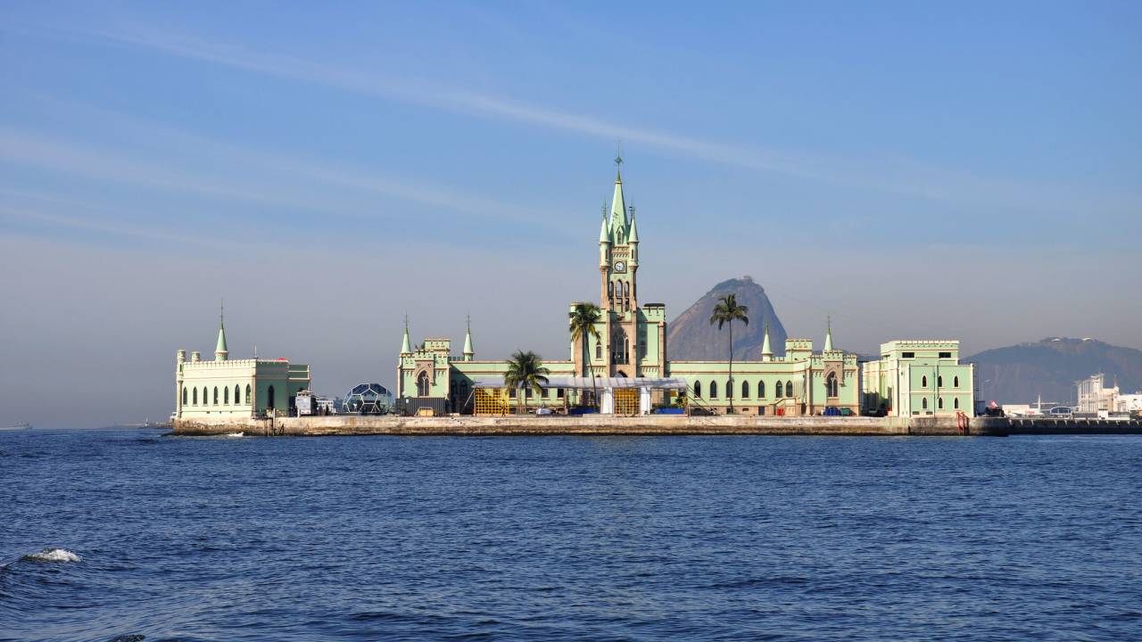 A Ilha Fiscal vista de longe, com o mar na frente, o palacete neogótico verde na filha e o Pão de Açúcar atrás