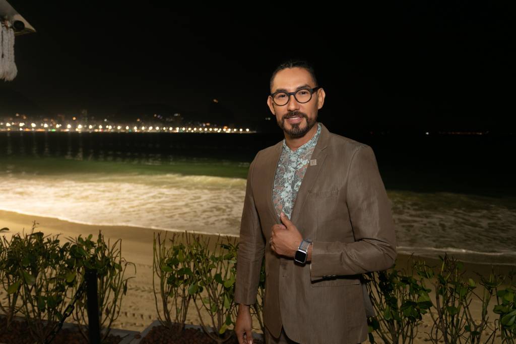 Foto mostra diretor corporativo usando paletó e blusa de renda com a praia de copacabana ao fundo