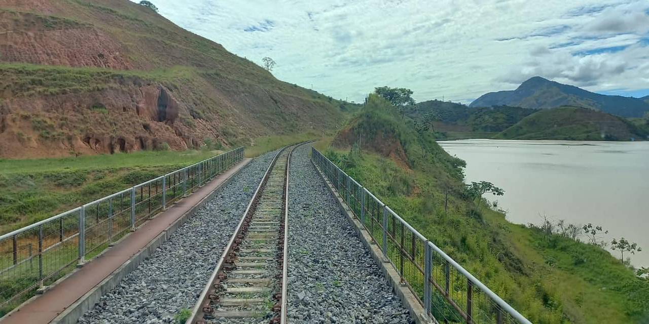 Foto mostra trilho de trem passando por paisagem. Há um Rio do lado direito da foto e uma montanha do lado esquerdo.
