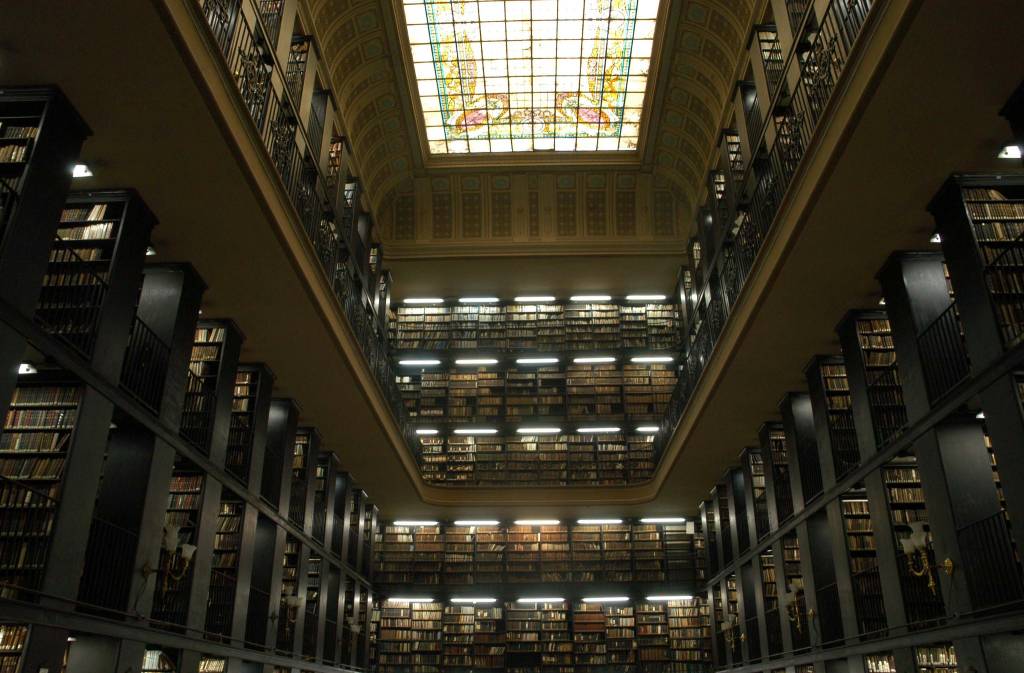 Foto interna da bibiloteca nacional, com um vitral retangular no teto e diversas plateleiras repletas de livros em três paredes, de cima a baixo, em dois pisos.