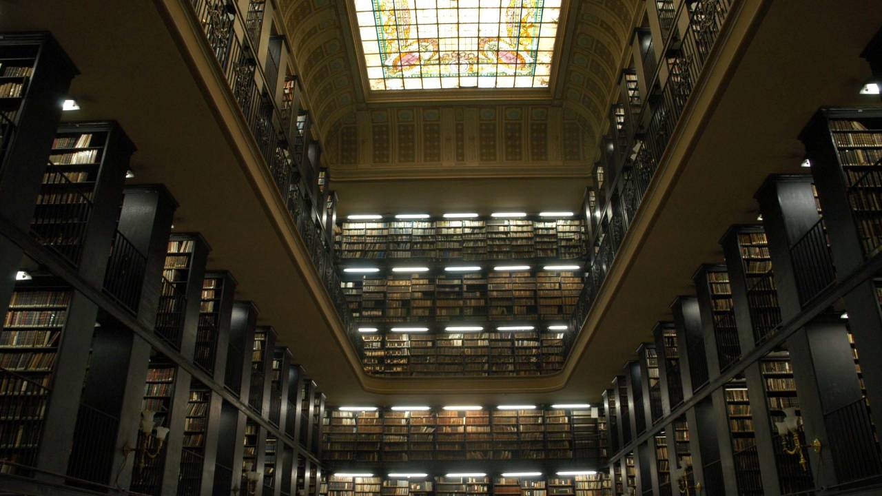 Foto interna da bibiloteca nacional, com um vitral retangular no teto e diversas plateleiras repletas de livros em três paredes, de cima a baixo, em dois pisos.