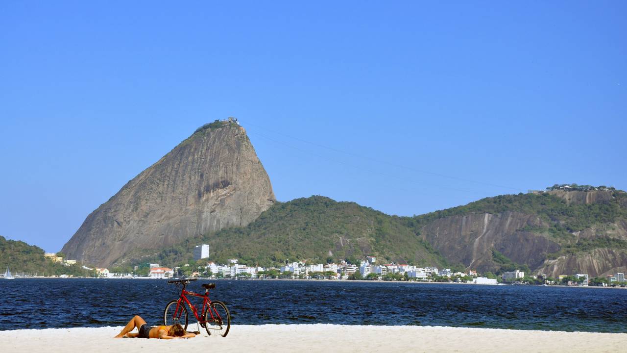 Homem deitado na areia da Praia do Flamengo, com uma biclecta vermelha de pé e o mar, o Morro da Urca e o Pão de Açúcar ao fundo.