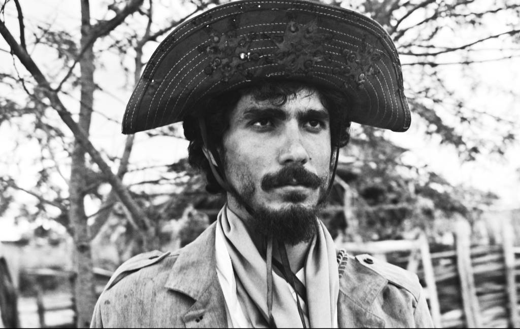 Frame do ator Vertin Moura, homem branco com cabelo, barba e bigode escuros, com chapéu de cangaceiro, em preto e branco