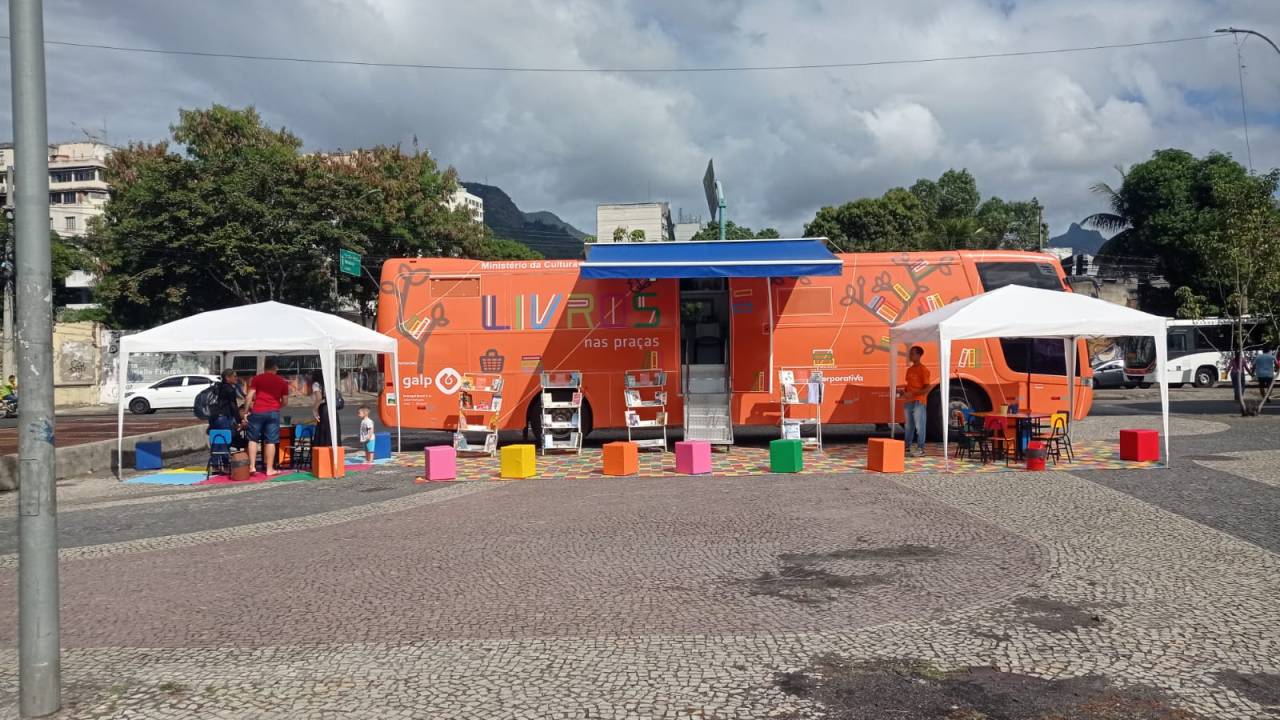 Praça com ônibus-biblioteca com de laranja e duas tendas brancas na frente, uma de cada lado