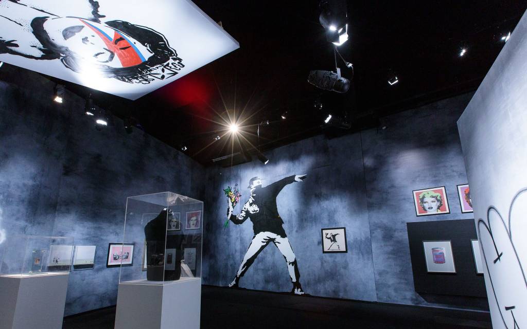 Sala de paredes cinza com obras de Banksy expostas, com destaque para Flower Thrower ao fundo (um homem se preparando para atirar um buquê de flores) e Queen Ziggy no teto (a rainha Elizabeth com a maquiagem que David Bowie usou na capa do disco Aladdin Sane, com o famoso raio vermelho e azul).
