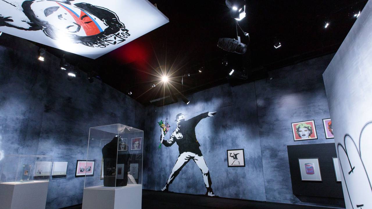 Sala de paredes cinza com obras de Banksy expostas, com destaque para Flower Thrower ao fundo (um homem se preparando para atirar um buquê de flores) e Queen Ziggy no teto (a rainha Elizabeth com a maquiagem que David Bowie usou na capa do disco Aladdin Sane, com o famoso raio vermelho e azul).