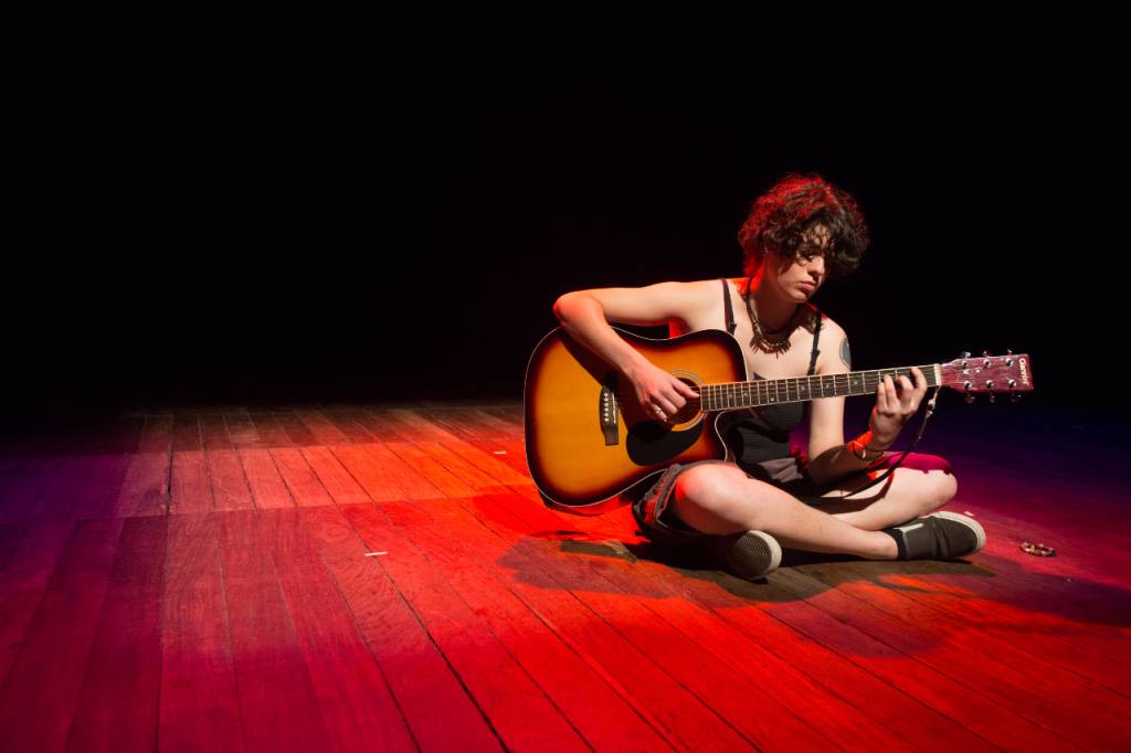 Tacy de Campos é uma mulher branca, com cabelos castanhos médios. Ela está sentada sobre um piso de madeira tocando violão. Usa bermuda e camiseta preta de alcinha.