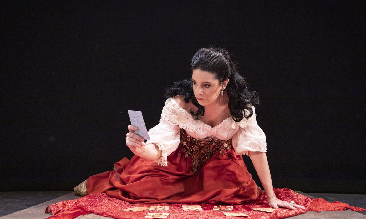 Mulher branca de cabelos pretos com saia vermelha e camisa branca, ajoelhada no chão, diante de uma toalha vermelha, consultando as cartas de um baralho.