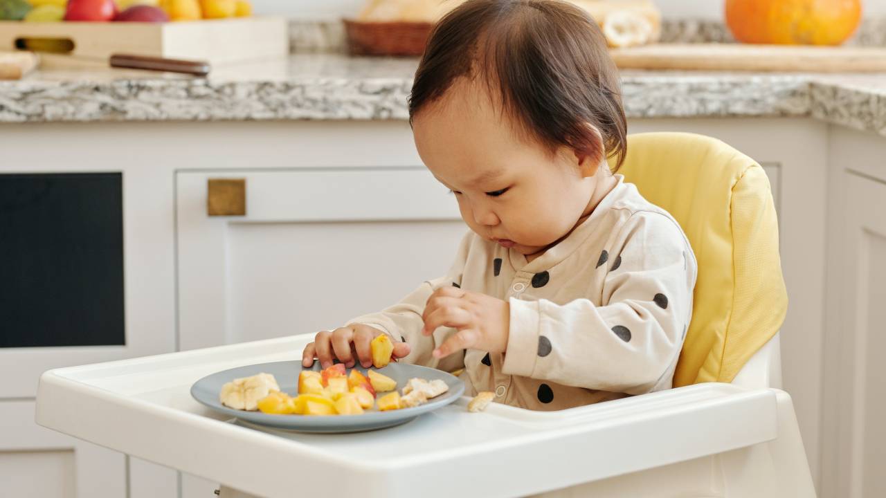 Bebê na cadeira de alimentação tentando comer frutas