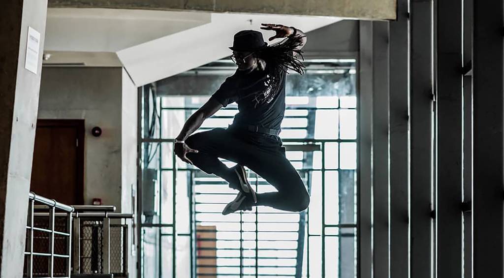 Silhueta do dançarino André Feijão saltando, com uma janela e suas grades ao fundo