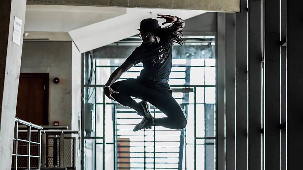 Silhueta do dançarino André Feijão saltando, com uma janela e suas grades ao fundo