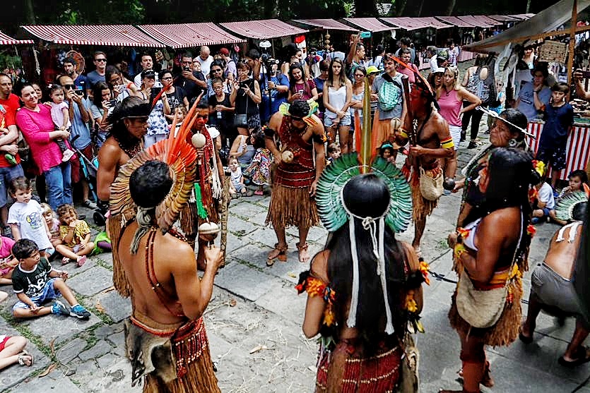 Indígenas em círculo são observados pelo público do evento. Ao fundo, barracas da feira do evento.
