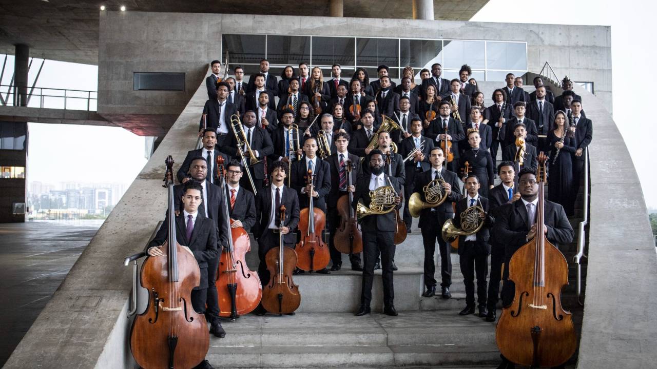 Os músicos estão de terno preto, com seus instrumentos, sobre uma escada de concreto