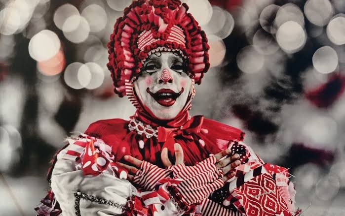 Foto em preto e branco de um homem com fantasia de carnaval quye lembra um clown, com detalhes em vermelho e em branco sobre a foto