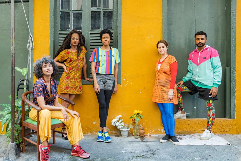 As cinco atrizes do elenco em frente a uma fachada amarela com porta e janela cinzas