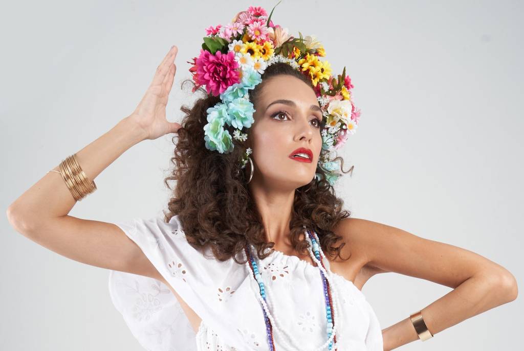 Bruna Pazinato é uma mulher branca de cabelo castanho cacheado, Ela usa vestido branco, arranjo de flores coloridas na cabeça e tem uma das mãos próxima à cabeça e a outra na cintura. Foto sobre fundo branco.
