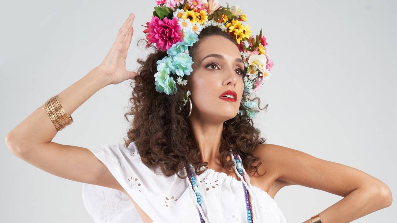 Bruna Pazinato é uma mulher branca de cabelo castanho cacheado, Ela usa vestido branco, arranjo de flores coloridas na cabeça e tem uma das mãos próxima à cabeça e a outra na cintura. Foto sobre fundo branco.