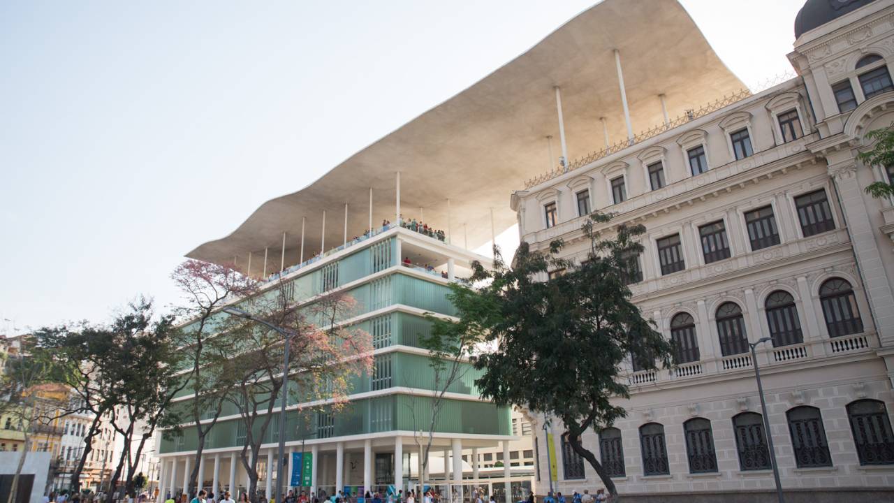 À esquerda, um prédio todo envidraçado, com pilotis brancos. À direita um edifício de época. Os dois fazem parte do Museu do Arte do Rio e são unidos por um teto ondulado branco