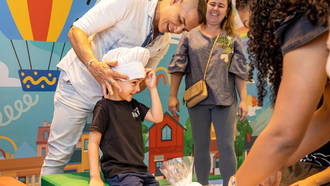 Foto mostra criança vestida como chef de cozinha, com pai colocando chapéu de cozinheiro em sua cabeça