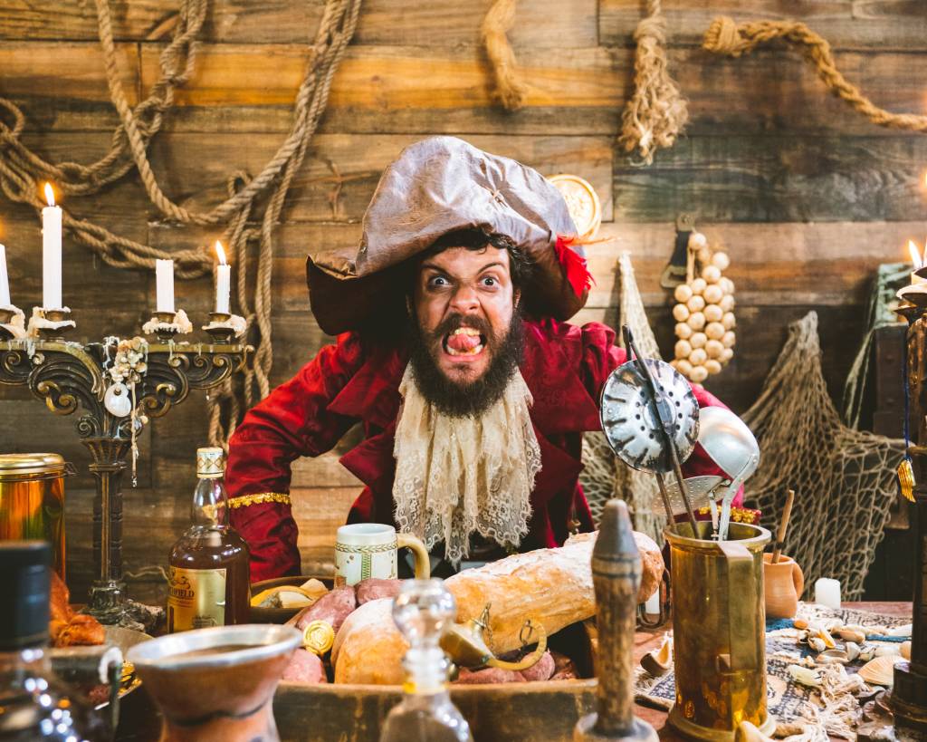 Diogo Defante é um homem branco com cabelo, barba e bigode castanhos, usando roupa de pirata com capa vinho e chapéu, diante de uma mesa cheia de objetos.