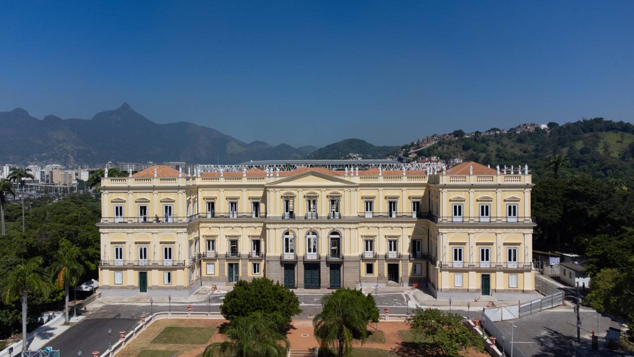 A fachada do palácio que abriga o Museu Nacional, em amarelo-claro, fotografada à distância, com o jardim à frente.