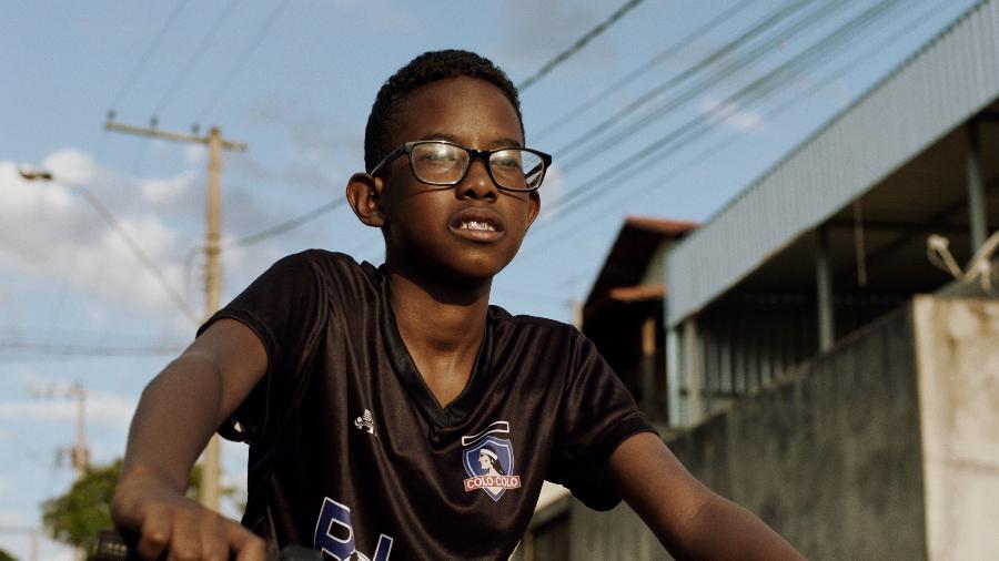 O ator Cícero Lucas é um menino negro, que está de cabelo curto e óculos de grau. Ele usa blusa preta com estampas e está montado em uma bicicleta